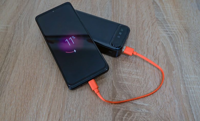 A smartphone charging from a power bank with an orange cable on a wooden table.