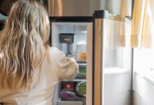 Back view of a blonde woman retrieving food from a refrigerator in a bright kitchen.