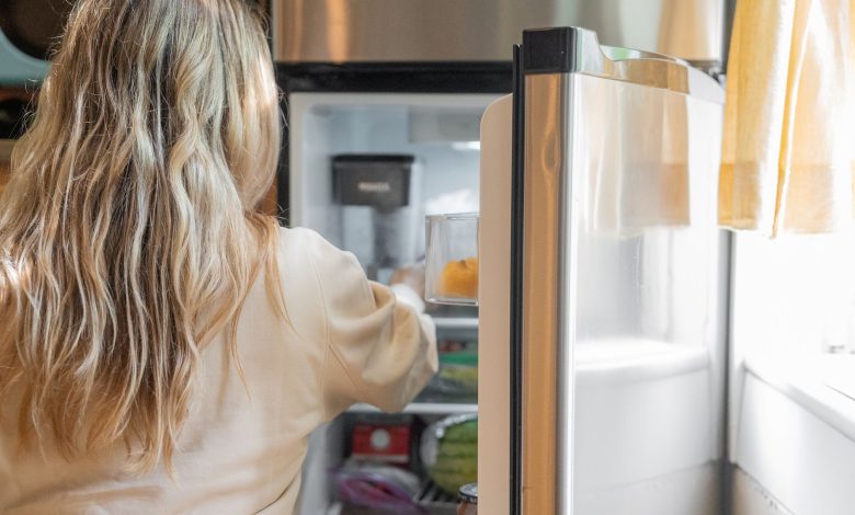 Back view of a blonde woman retrieving food from a refrigerator in a bright kitchen.