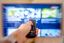 Hand holding remote control pointing at a blurred TV screen in a cozy indoor setting.