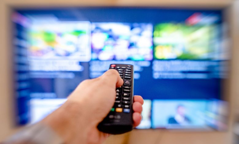 Hand holding remote control pointing at a blurred TV screen in a cozy indoor setting.