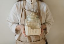 A close-up of hands holding a glass jar filled with flour, wearing a beige apron.