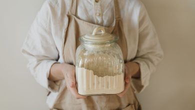 A close-up of hands holding a glass jar filled with flour, wearing a beige apron.
